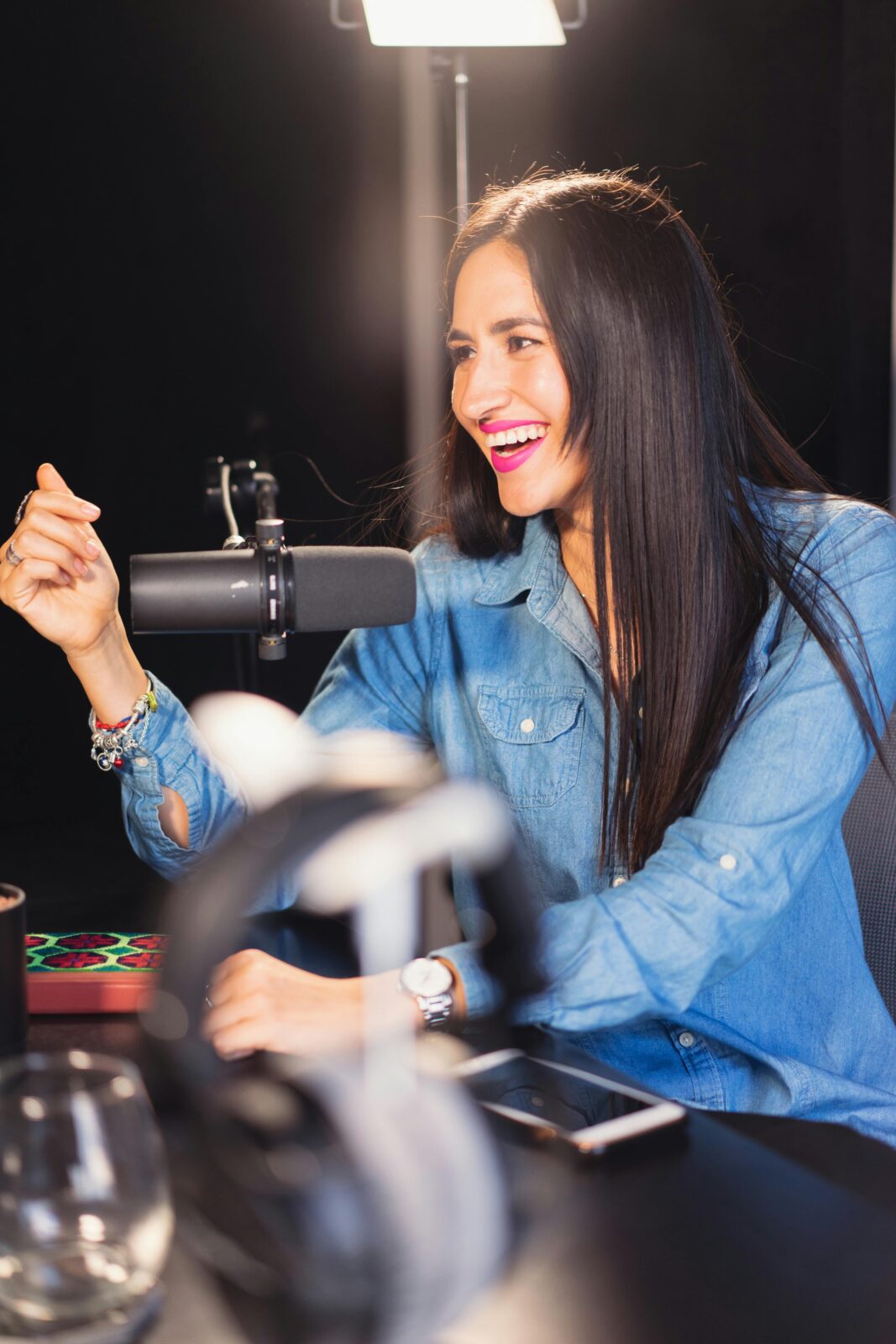 Smiling woman in denim recording a lively podcast in a professional studio setting.