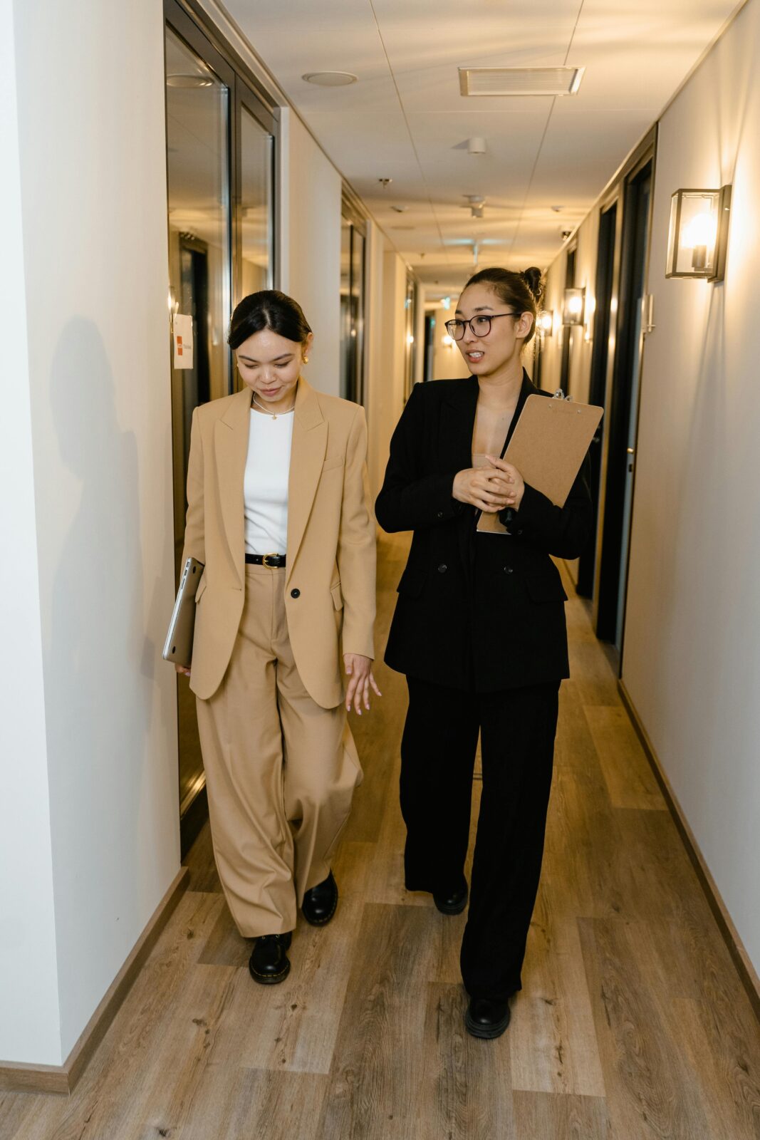 Two professional women walking in an office hallway, discussing work while carrying documents.