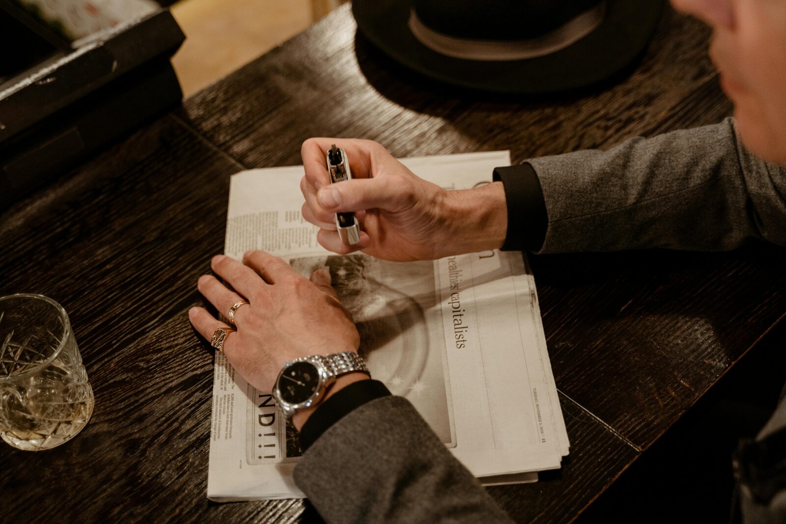 Man in formal attire reading newspaper, holding pen, and wearing watch at stylish desk.